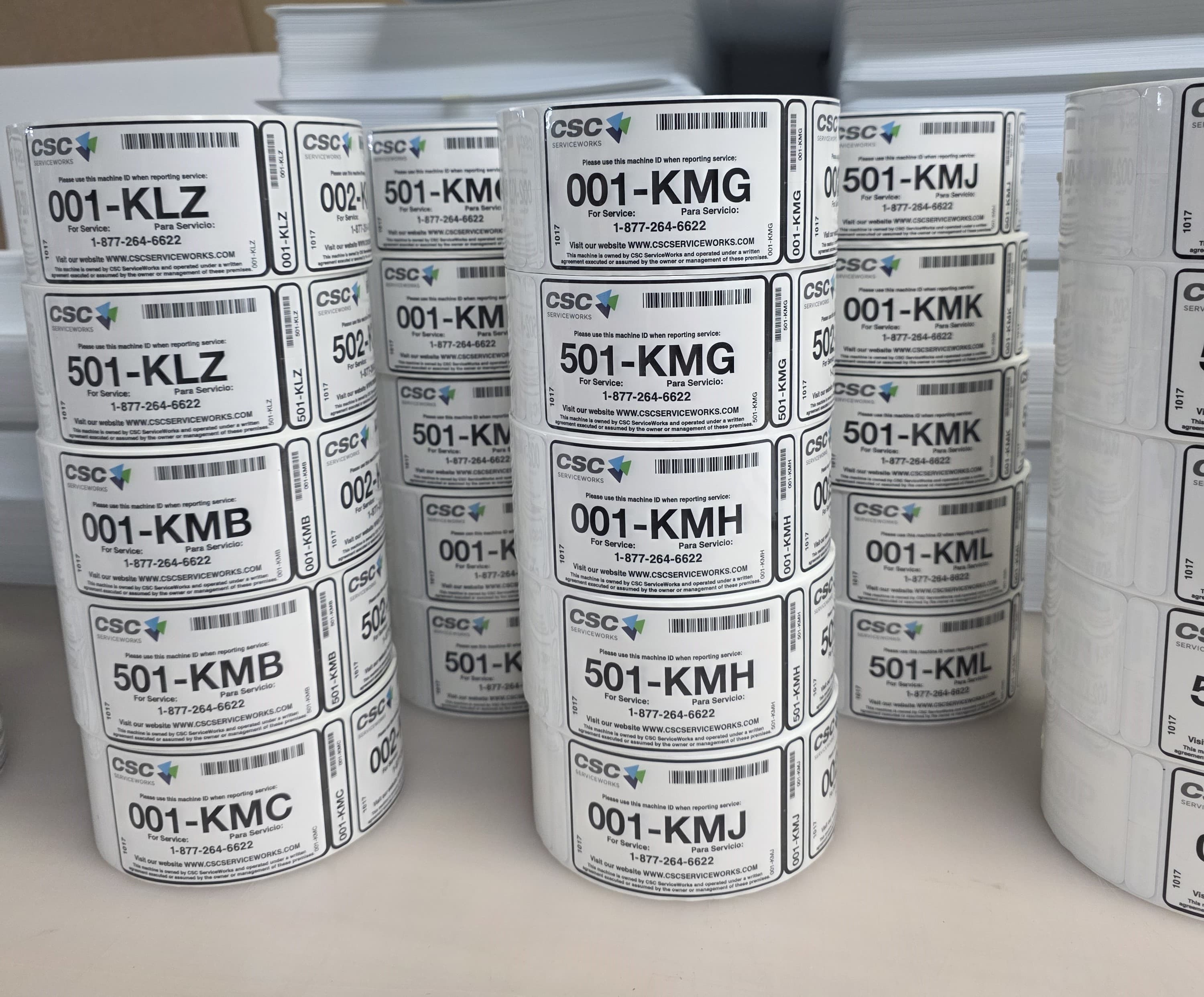 Stacked rolls of white service labels, each carrying a barcode, a unique alphanumeric asset code and bilingual service instructions