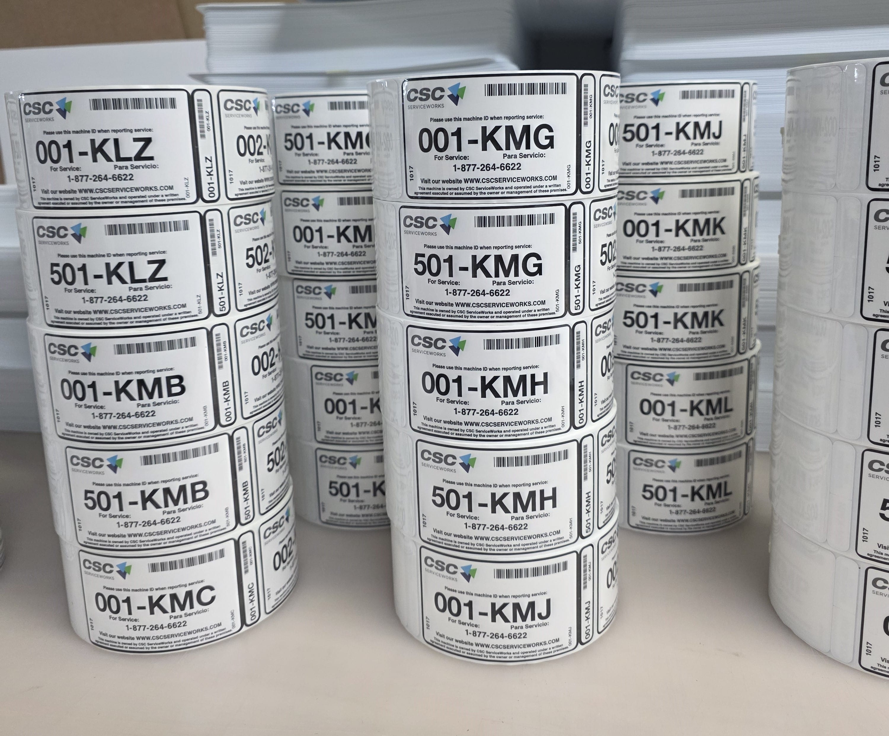 Stacked rolls of white service labels, each carrying a barcode, a unique alphanumeric asset code and bilingual service instructions