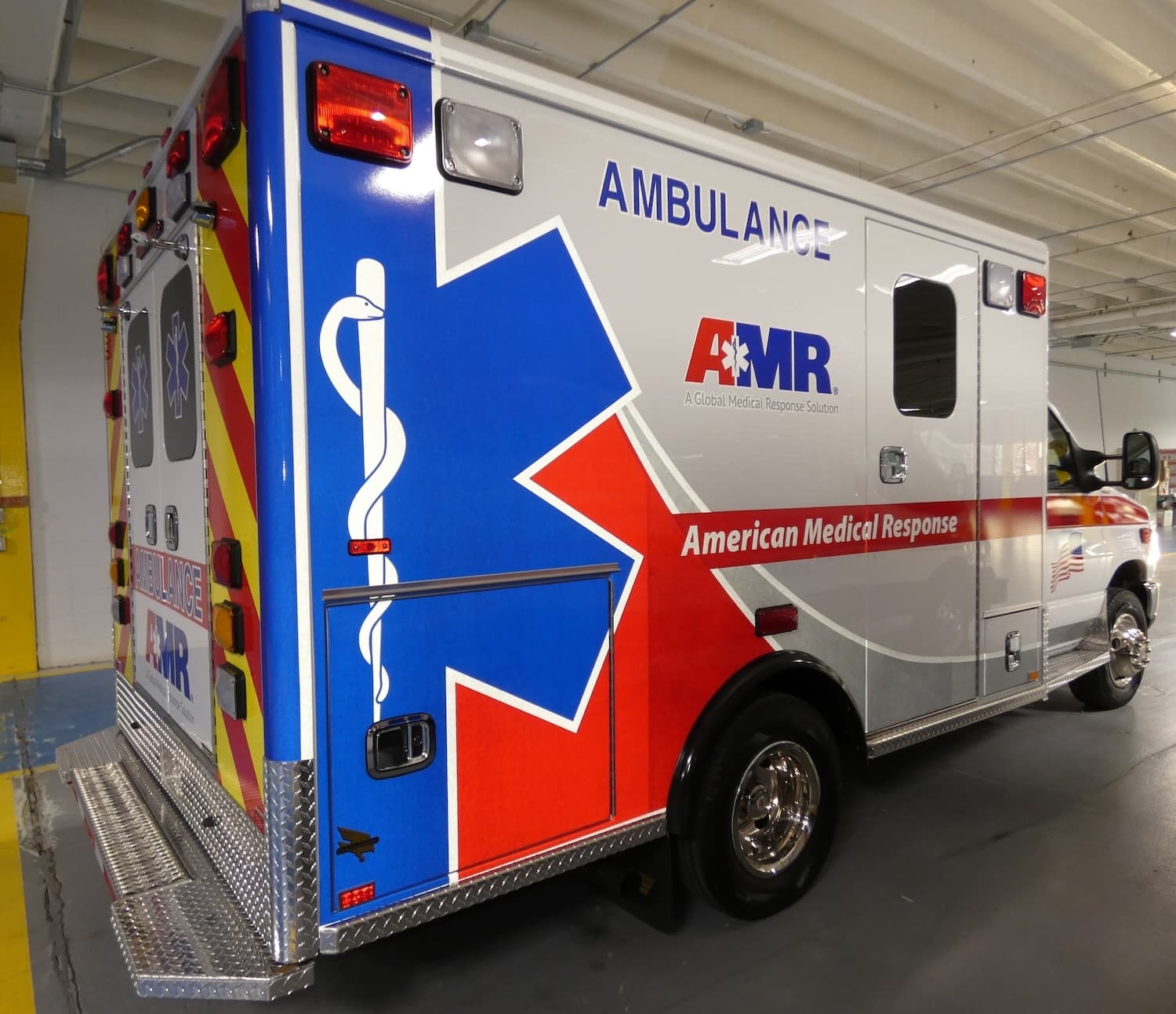 Rear three-quarter view of a white AMR ambulance with a large blue and red star of life, AMBULANCE lettering, a red American Medical Response stripe and red and yellow chevrons around the rear doors