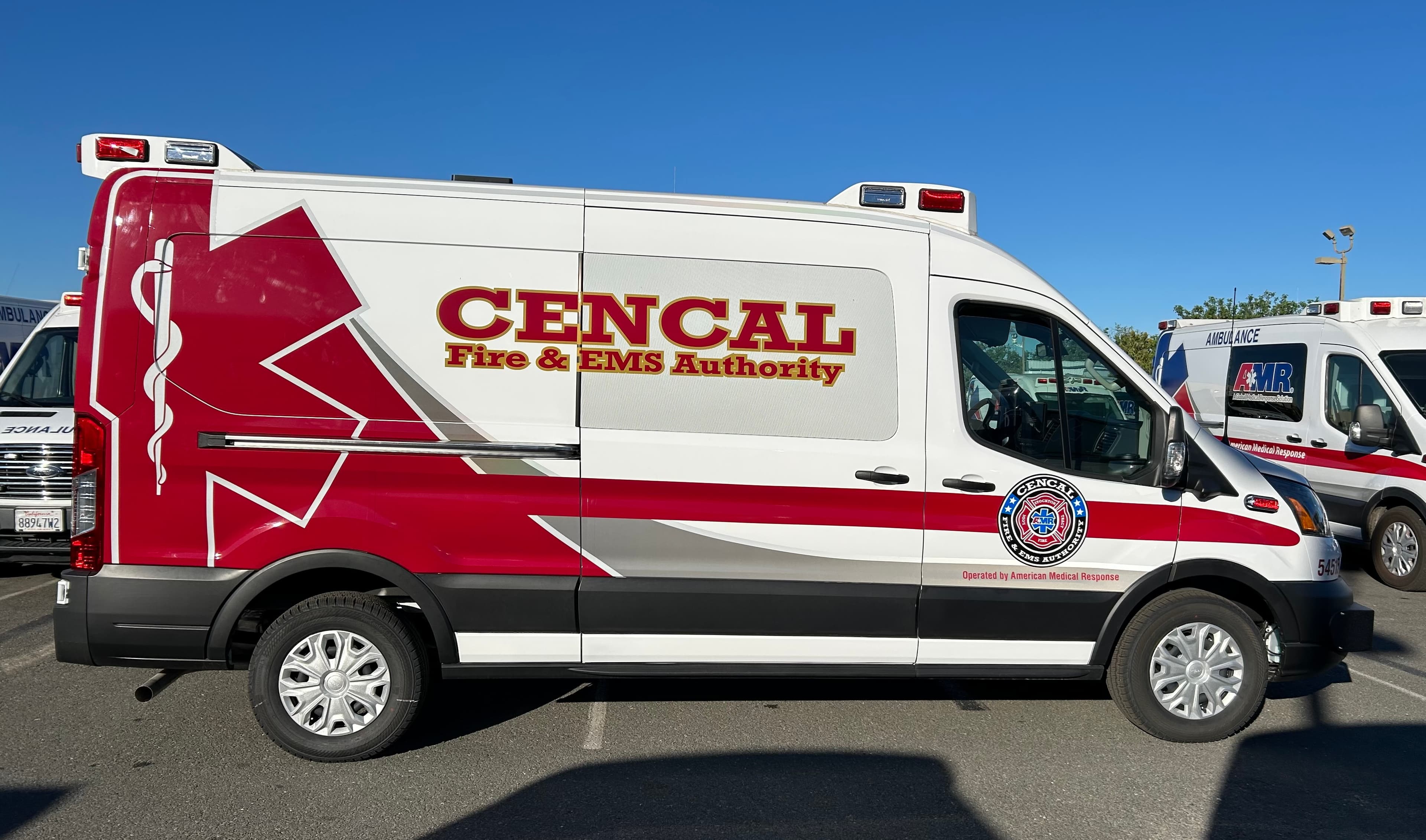 White high-roof ambulance van with CENCAL Fire & EMS Authority lettering in red and gold, a large red star of life across the rear panels and the authority seal on the passenger door, parked beside AMR ambulances